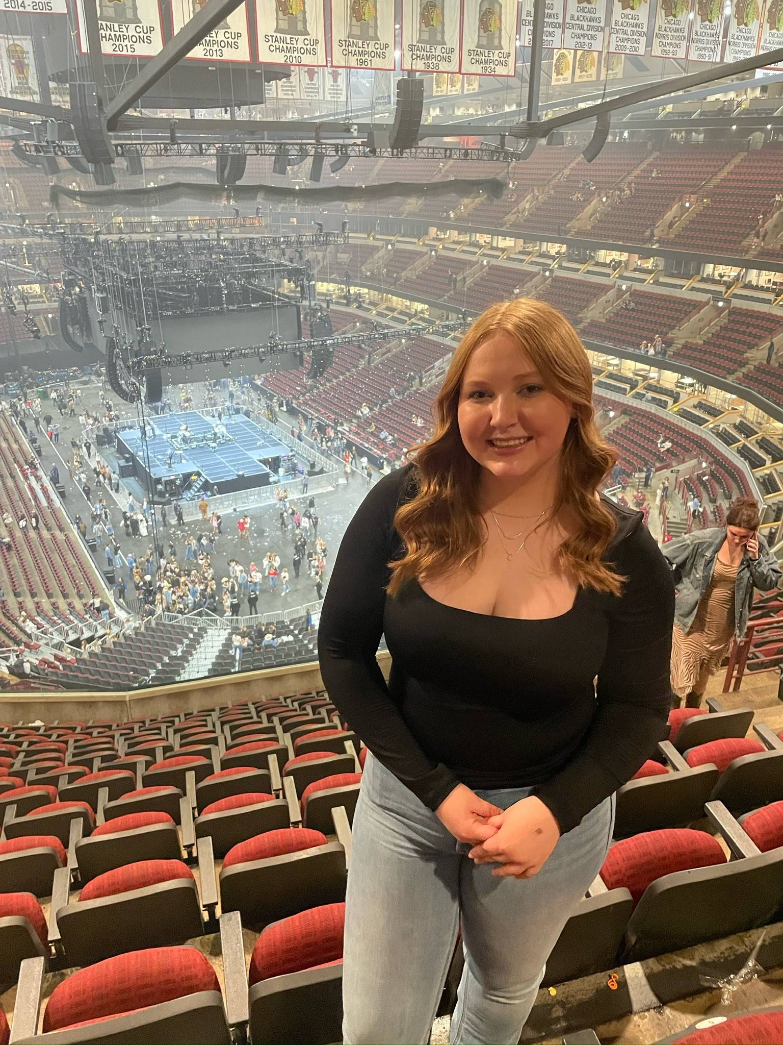 Young woman standing in a nearly empty arena with the stage and balcony seats in the background. She has curly hair and is wearing a black shirt with light colored jeans.